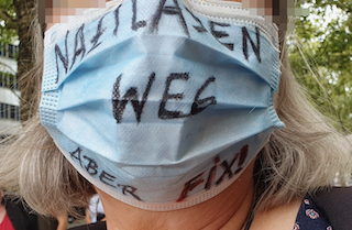 Foto zeigt: Gesicht mit hellblauer Schutzmaske; darauf die Aufschrift 'Naziladen weg aber fix'