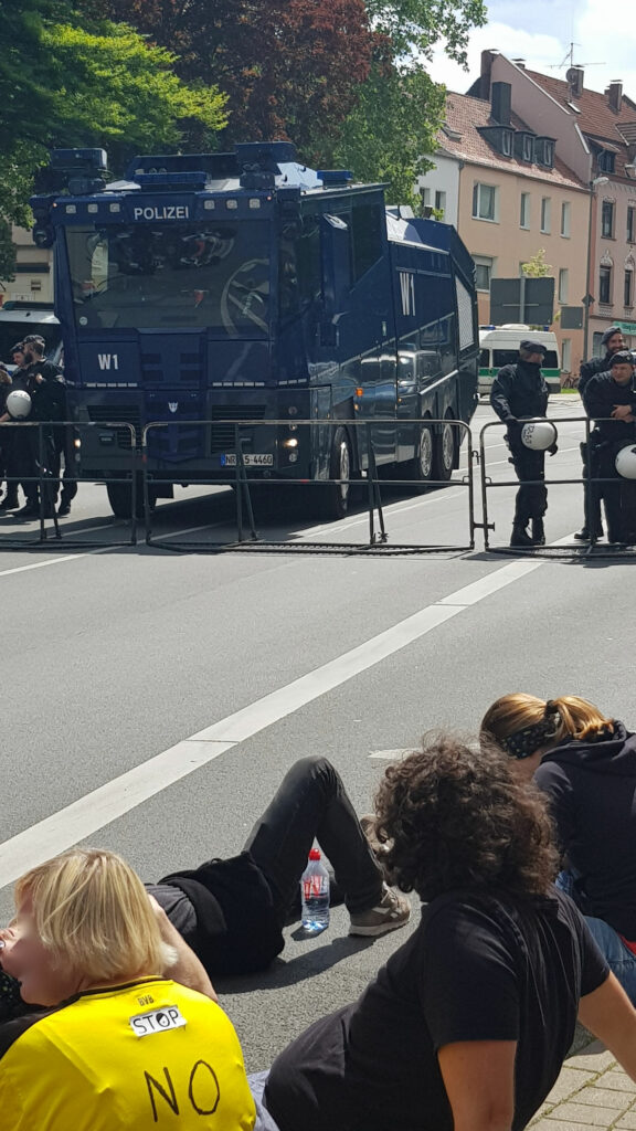 Foto zeigt: Menschen sitzen und liegen auf der Straße, im Hintergrund eine Absperrung,   ein Wasserwerfer und Polizist*innen